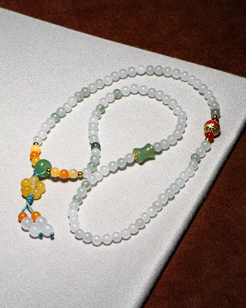 A beaded necklace with white, green, yellow, and orange beads, featuring a cluster of yellow beads and a decorative tassel, displayed on a white and brown textured surface.