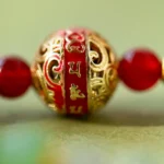 A close-up of a decorative bead with intricate gold patterns and red detailing, featuring small embossed symbols or characters on a red band, flanked by two red beads on either side.