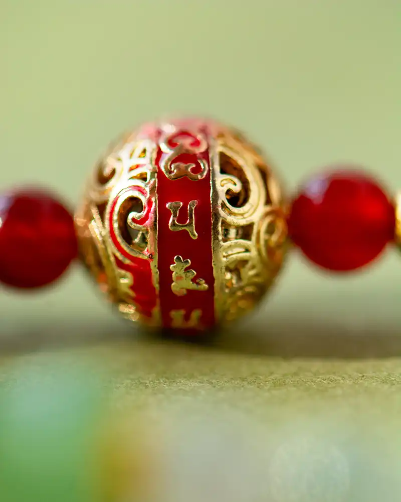 A close-up of a decorative bead with intricate gold patterns and red detailing, featuring small embossed symbols or characters on a red band, flanked by two red beads on either side.