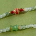 Close-up of a beaded necklace with pale green round beads, a green barrel-shaped bead, red beads, gold accents, and a decorative gold and red bead with Chinese characters, on a green background.