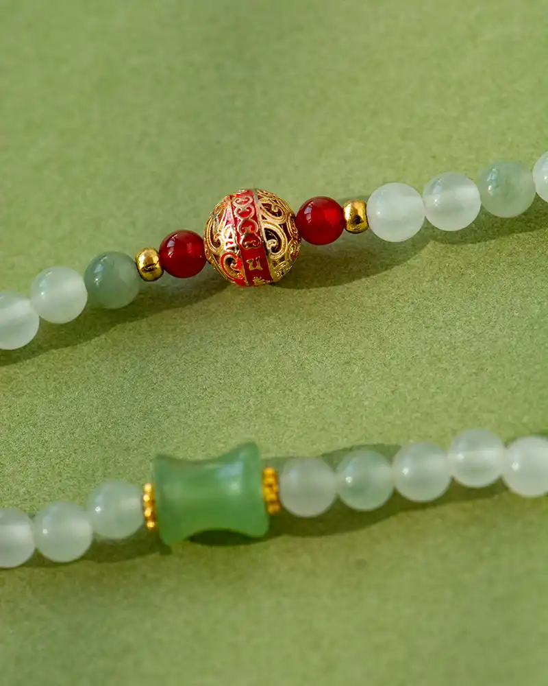 Close-up of a beaded necklace with pale green round beads, a green barrel-shaped bead, red beads, gold accents, and a decorative gold and red bead with Chinese characters, on a green background.