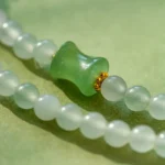 A close-up of a beaded necklace with smooth, round white beads and a central green bead accented with small gold details, set against a soft green background.