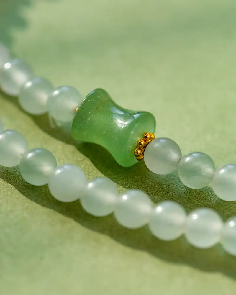 A close-up of a beaded necklace with smooth, round white beads and a central green bead accented with small gold details, set against a soft green background.