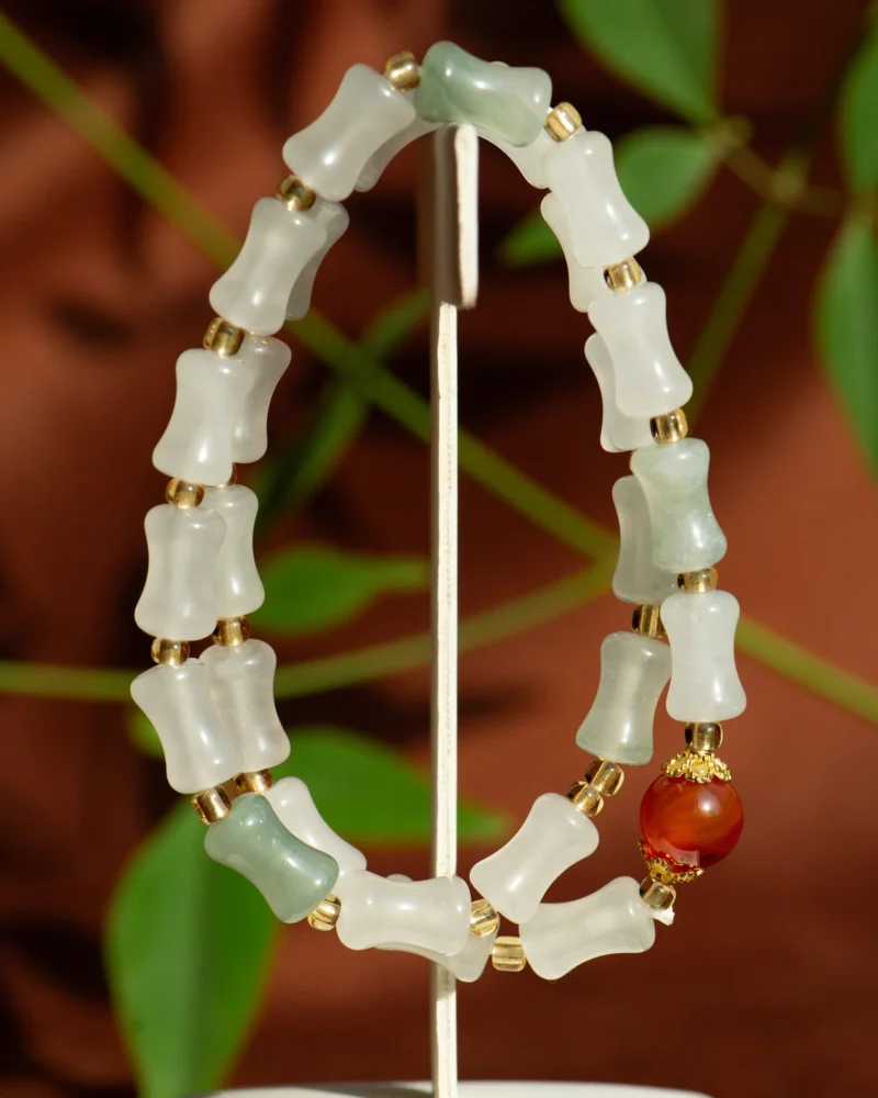 The White and Light Green Quartzite Jade Beaded Adornment—featuring delicately carved links, accented with gold spacers and a bold red bead—rests elegantly on a stand against a softly blurred backdrop of brown and green foliage.