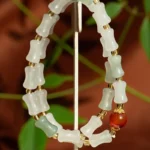 A beaded bracelet with frosted, bone-shaped white beads, green accent beads, small gold spacers, and a single round amber bead displayed on a stand with green leaves in the background.