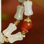 A close-up of a beaded bracelet featuring frosted white, bone-shaped beads, small gold spacers, and a single round red bead with ornate gold caps against a blurred background.