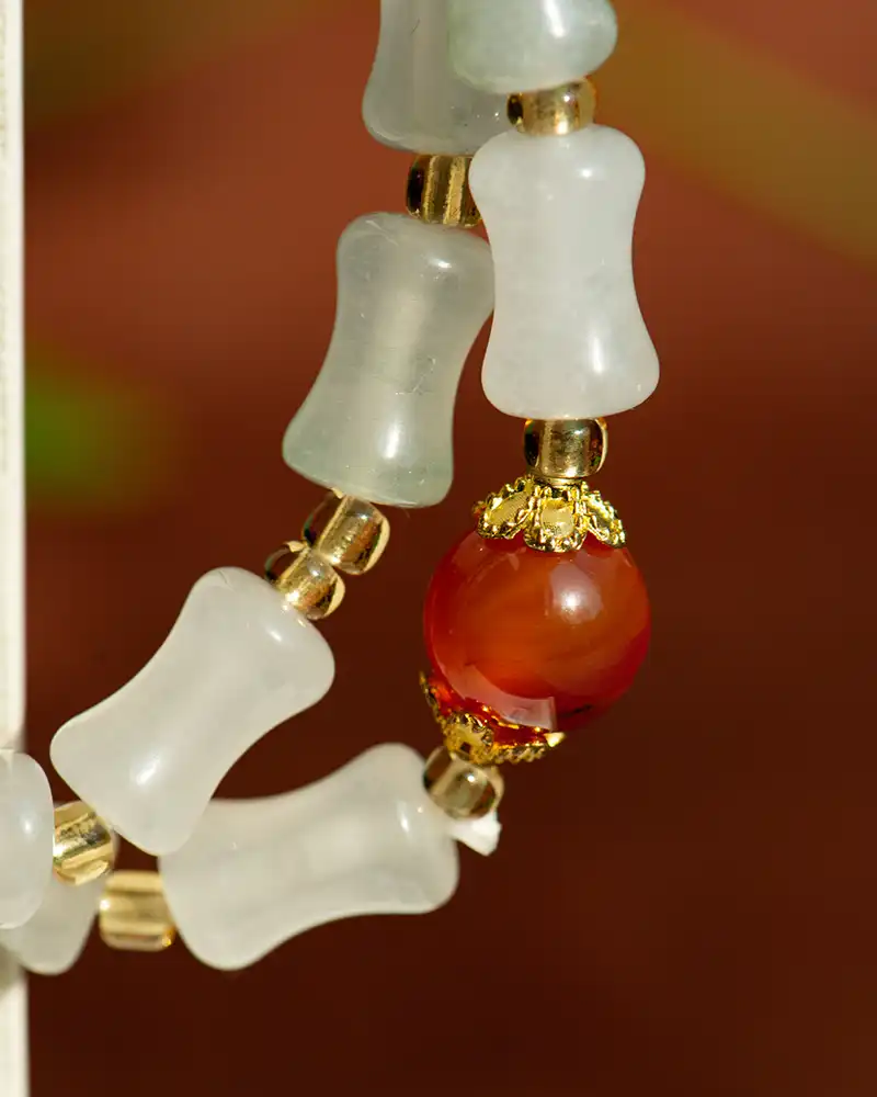 LYU_4820 A close-up of a beaded bracelet featuring frosted white, bone-shaped beads, small gold spacers, and a single round red bead with ornate gold caps against a blurred background.