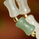 Close-up of a beaded necklace with translucent white and greenish barrel-shaped beads, separated by small gold round beads, set against a blurred brown background.
