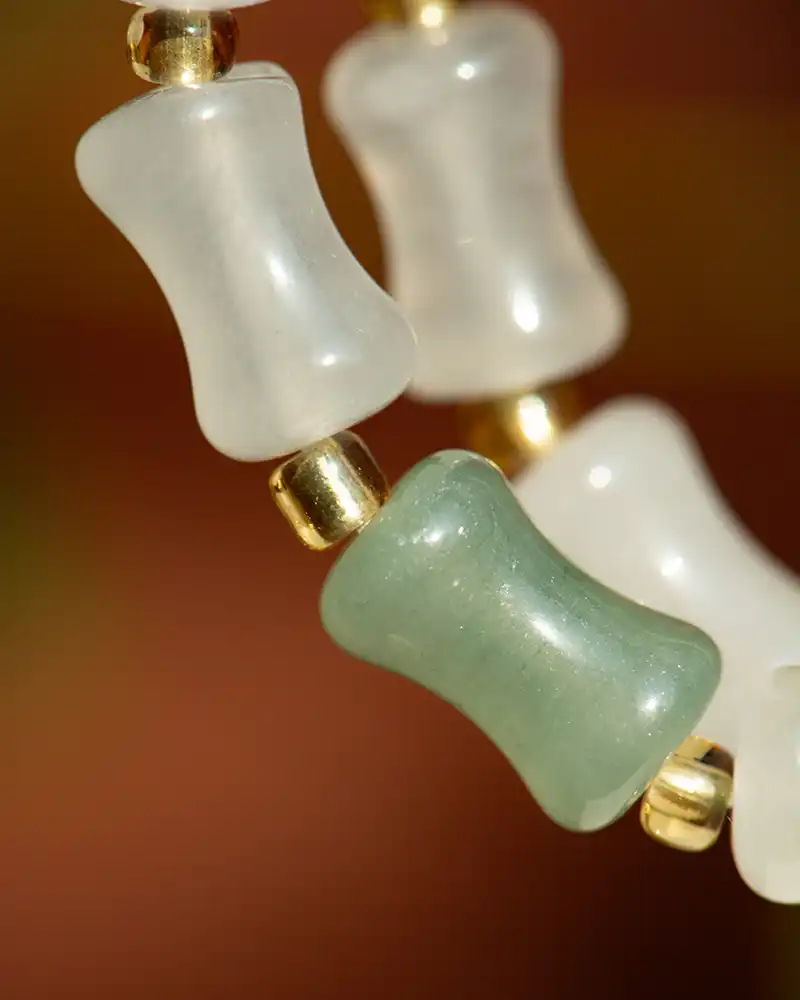 LYU_4821 Close-up of a beaded necklace with translucent white and greenish barrel-shaped beads, separated by small gold round beads, set against a blurred brown background.