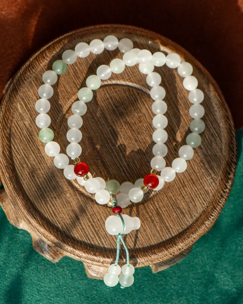 The White Quartzite Jade Beaded Bracelet, featuring red accents and a double-drop detail, is showcased coiled on a round wooden platform against a green and brown background.