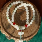 A beaded necklace with white and green beads, accented by a few red beads, is arranged in a circle on a round wooden surface with a green and brown background.