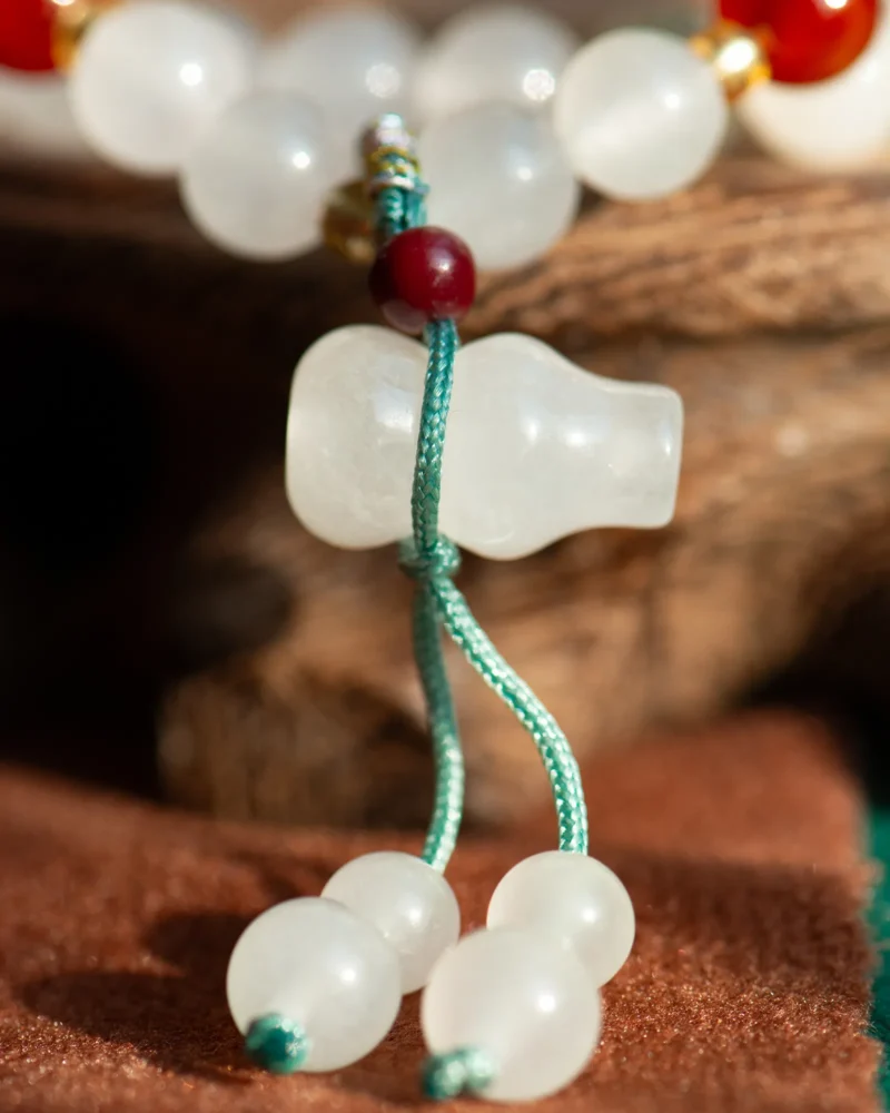 Close-up of the White Quartzite Jade Beaded Bracelet, showcasing white spherical beads, a larger double-drop bead, a red accent, and green string, set against a blurred wooden background.