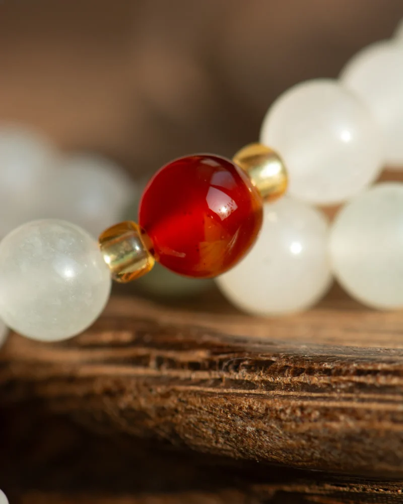 A close-up of the White Quartzite Jade Beaded Bracelet - Red Accent and Double-Drop Detail, showcasing its white translucent quartzite jade beads, a central shiny red accent bead, and delicate gold spacers, elegantly displayed on a textured brown surface.