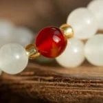 A close-up of a beaded bracelet featuring a single shiny red bead flanked by small gold beads, with larger frosted white beads in the background resting on a textured wooden surface.