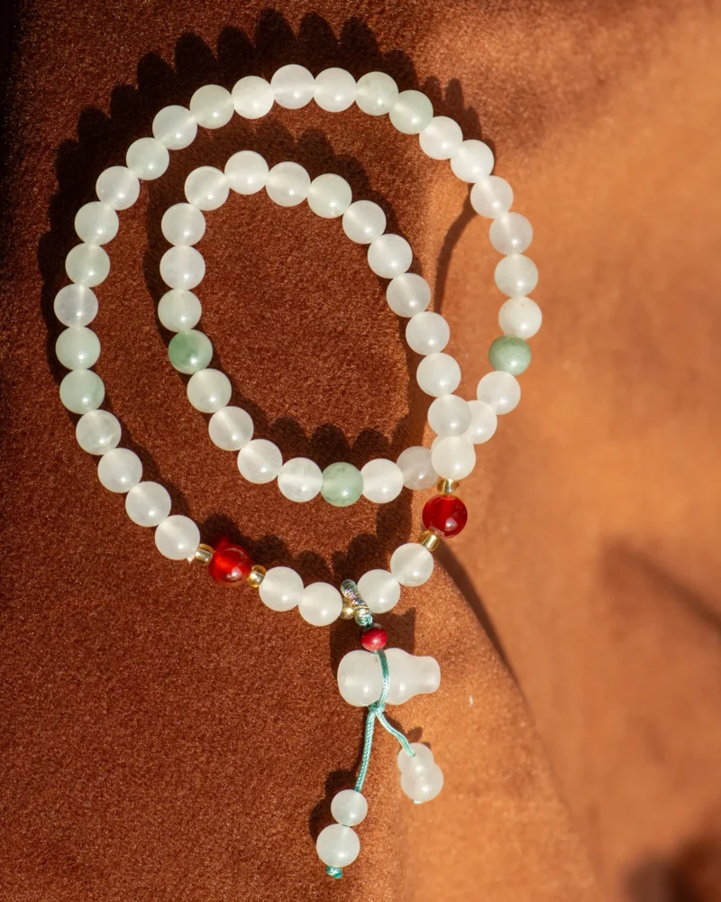 Certainly! Here’s the rewritten sentence using your product data: The White Quartzite Jade Beaded Bracelet features delicate white beads accented with pale green and red, artfully arranged to showcase the subtle hues characteristic of quartzite jade jewelry. Finished with a blue string and double-drop detail, this bracelet’s dangling beads add a charming touch reminiscent of elegant craftsmanship.