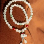 A beaded necklace with white, green, and red beads, arranged in a spiral on a brown surface. The necklace features a blue string and a pendant with dangling beads at the bottom.