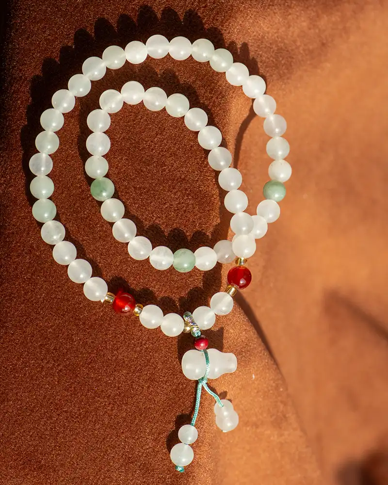 A beaded necklace with white, green, and red beads, arranged in a spiral on a brown surface. The necklace features a blue string and a pendant with dangling beads at the bottom.