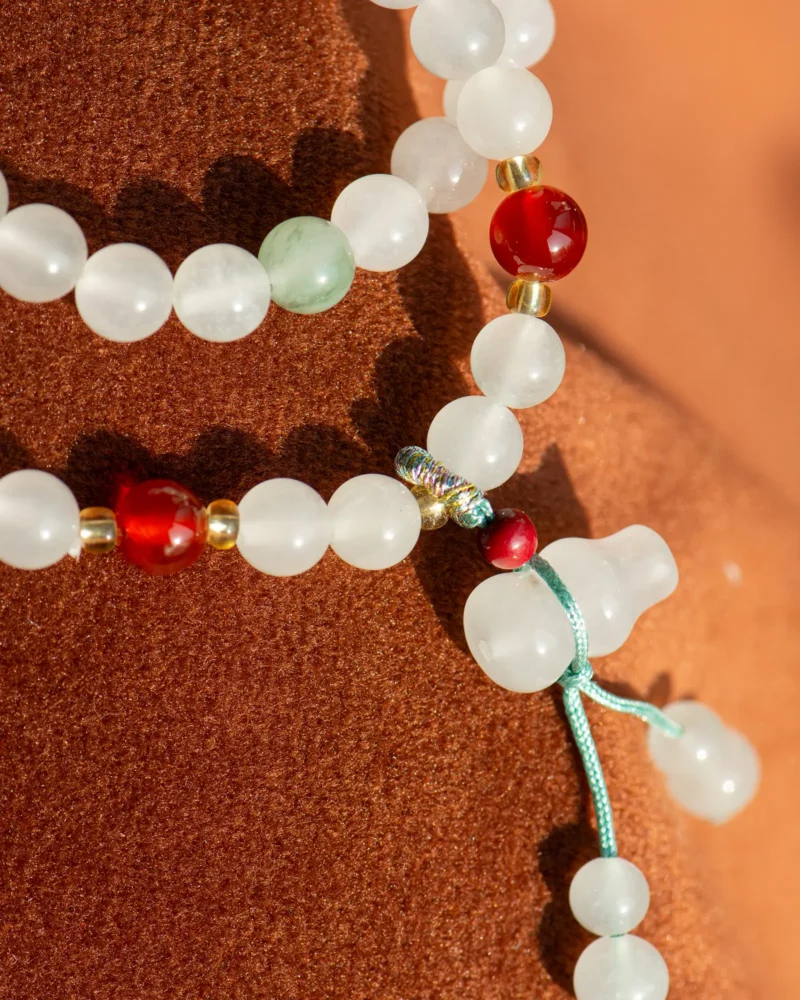 Close-up of the White Quartzite Jade Beaded Bracelet featuring white quartzite jade beads, red accent beads, a double-drop detail, and gold accents on a turquoise cord, displayed on textured brown fabric.