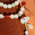 Close-up of a beaded necklace with white, red, and green beads, gold accents, and a turquoise string, displayed against a textured brown background.