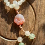A close-up of a beaded necklace with frosted white and green beads, featuring a central pink and white carved bead, displayed on a wooden surface with circular patterns.