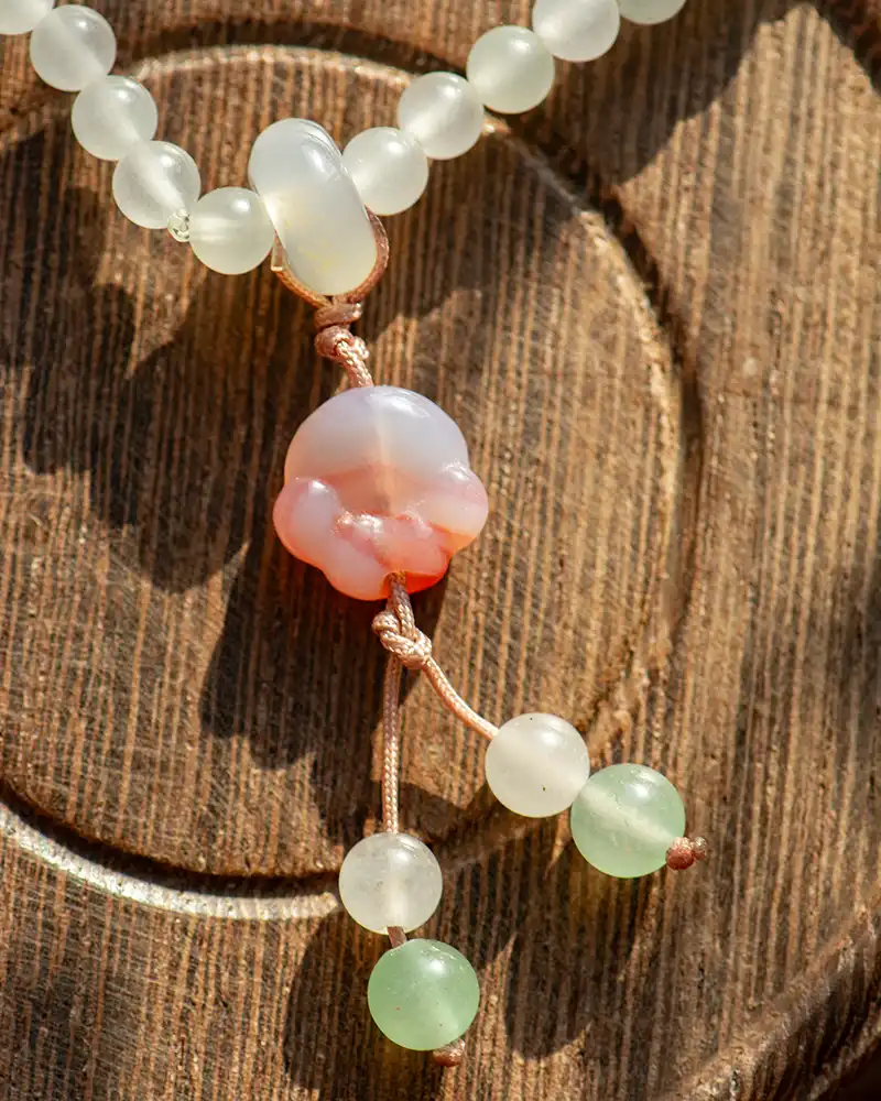 LYU_4829 A close-up of a beaded necklace with frosted white and green beads, featuring a central pink and white carved bead, displayed on a wooden surface with circular patterns.