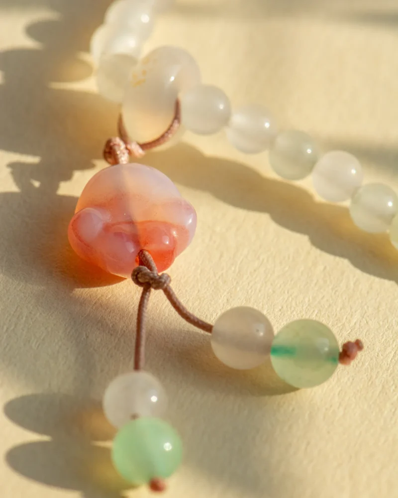 Close-up of the Quartzite Jade Bracelet with Carved Accents, showcasing frosted round beads and a central pinkish translucent paw-shaped bead, all strung on a brown cord and resting on a light surface in gentle sunlight.