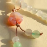 Close-up of a beaded necklace with frosted white and pale green round beads, featuring a translucent pink and orange carved bead resembling a cat’s face, all strung on light brown cord, lying on a light surface.