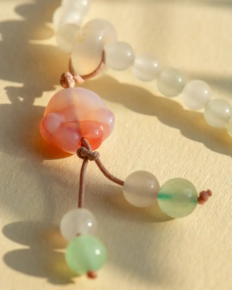 LYU_4831 Close-up of a beaded necklace with frosted white and pale green round beads, featuring a translucent pink and orange carved bead resembling a cat’s face, all strung on light brown cord, lying on a light surface.