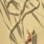 A beaded necklace with translucent white and pale green beads is arranged in a loose loop on a beige surface, ending in a pink seashell pendant with small green and white beads. Shadows from plants fall across the scene.
