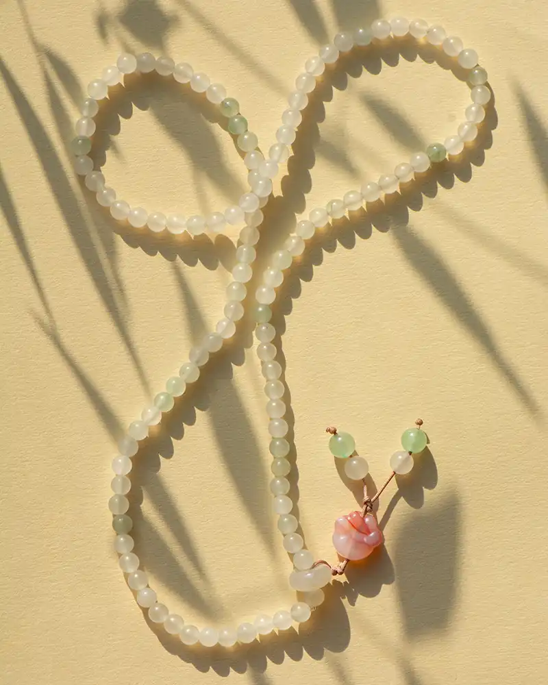 LYU_4833 A beaded necklace with translucent white and pale green beads is arranged in a loose loop on a beige surface, ending in a pink seashell pendant with small green and white beads. Shadows from plants fall across the scene.