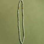 A long beaded necklace with small white beads, two orange beads, and green tassels at the ends, arranged on a plain, greenish background.