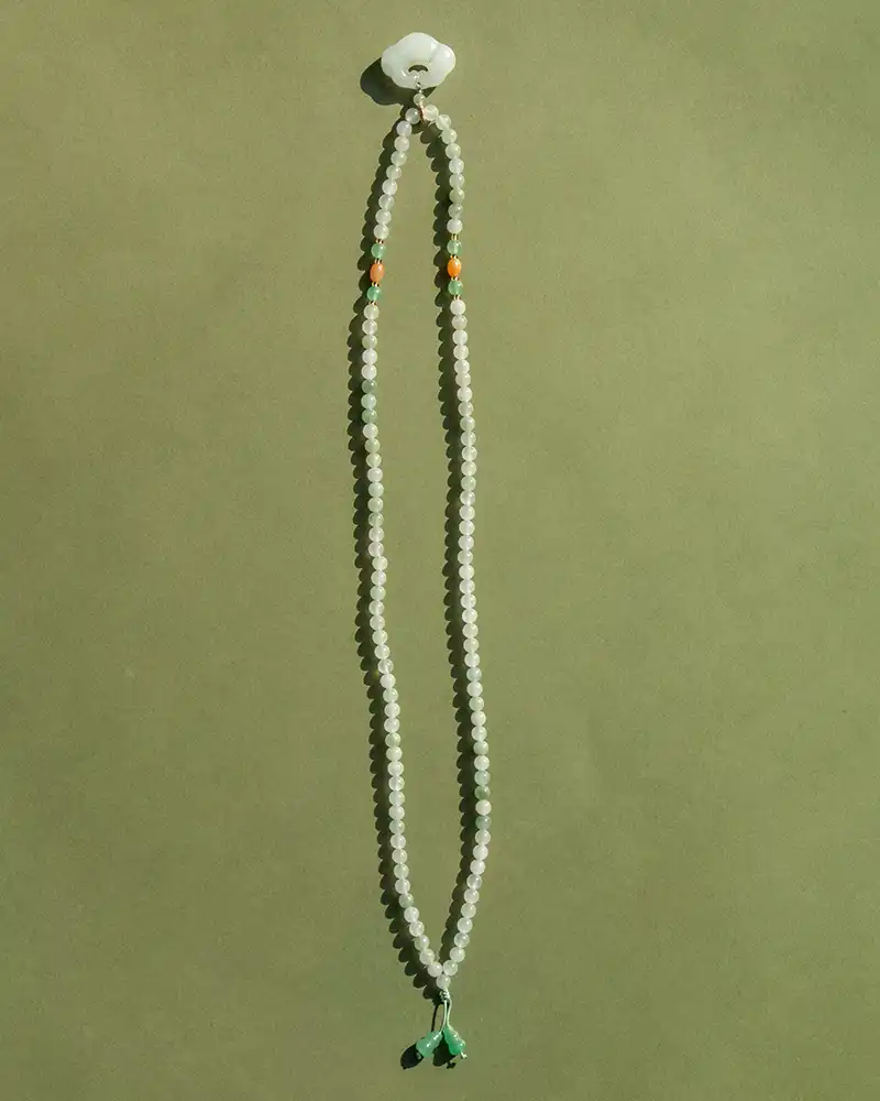 A long beaded necklace with small white beads, two orange beads, and green tassels at the ends, arranged on a plain, greenish background.