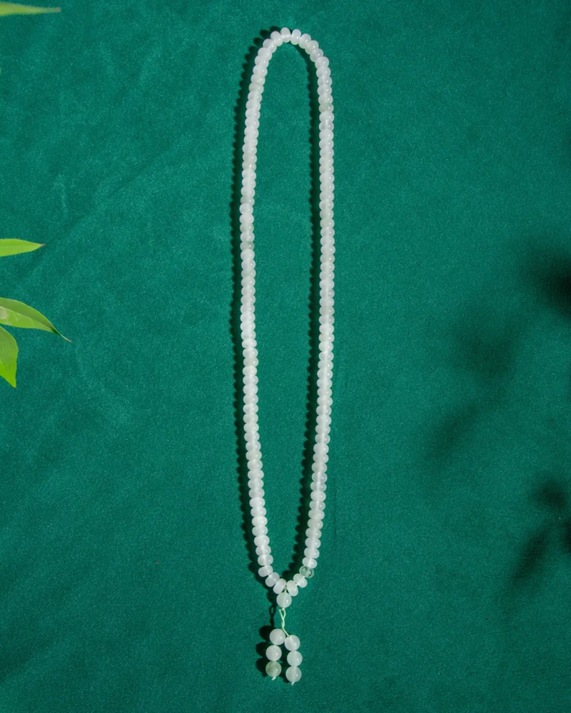 A White and Light Green Quartzite Jade Beaded Bracelet featuring a ring detail and twin drops is arranged in an oval shape on a green textured background, with a few green leaves visible at the edge.