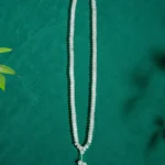 A long beaded white necklace with a pendant made of five round beads, displayed on a green textured background with a few green leaves visible in the corner.