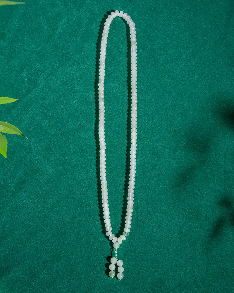 LYU_4856 A long beaded white necklace with a pendant made of five round beads, displayed on a green textured background with a few green leaves visible in the corner.
