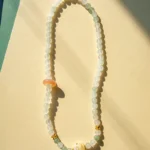 A beaded necklace with round, milky white and pale green beads, a larger pink bead, a barrel-shaped white bead with yellow patterns, and small gold accents, displayed on a light beige surface with soft shadows.
