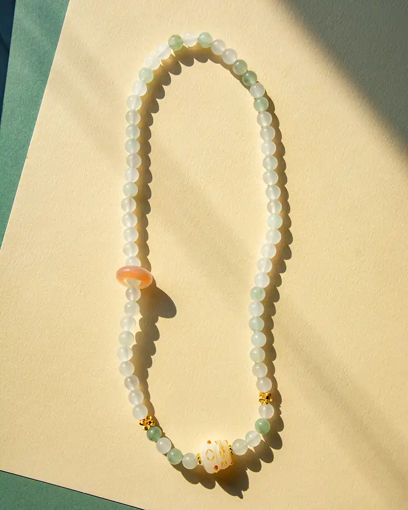 A beaded necklace with round, milky white and pale green beads, a larger pink bead, a barrel-shaped white bead with yellow patterns, and small gold accents, displayed on a light beige surface with soft shadows.