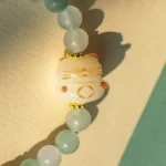 A close-up of a beaded bracelet featuring pale green round beads and a central bead shaped like a lucky cat, with gold accents, lying on a textured surface.