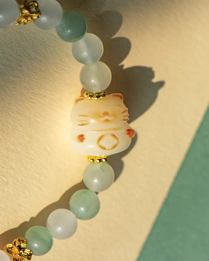 A close-up of a beaded bracelet featuring pale green round beads and a central bead shaped like a lucky cat, with gold accents, lying on a textured surface.