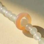 A close-up of a beaded necklace with translucent white round beads and one larger, peach-colored ring-shaped bead on a light beige background.