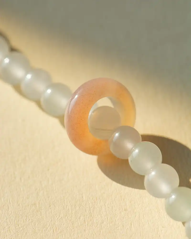 A close-up of a beaded necklace with translucent white round beads and one larger, peach-colored ring-shaped bead on a light beige background.