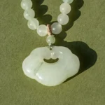 A jade beaded necklace with a round, cloud-shaped jade pendant rests on an olive-green surface, casting a soft shadow.