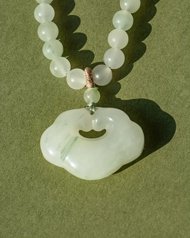 A jade beaded necklace with a round, cloud-shaped jade pendant rests on an olive-green surface, casting a soft shadow.