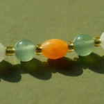 A close-up of a beaded necklace featuring green, white, and gold round beads with a prominent orange oval bead in the center, set against a light green background.