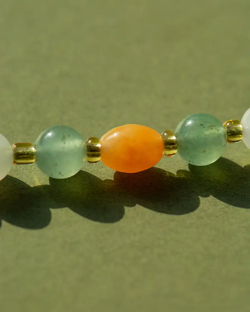 A close-up of a beaded necklace featuring green, white, and gold round beads with a prominent orange oval bead in the center, set against a light green background.