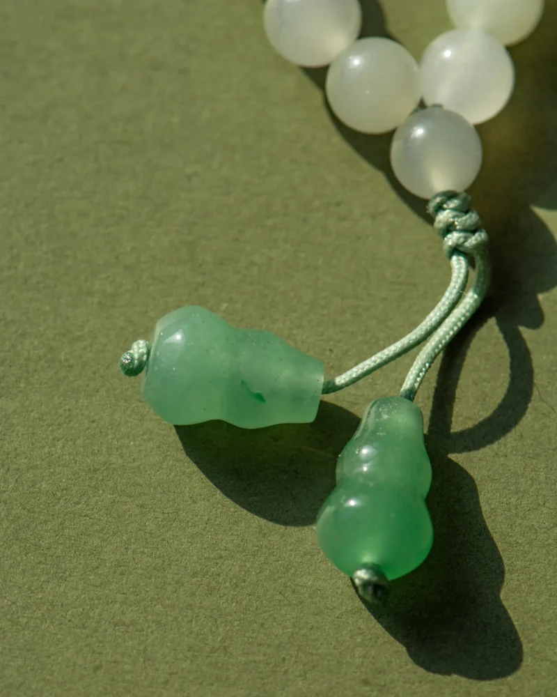 Close-up of the White and Light Green Quartzite Jade Necklace, featuring milky white round beads with a donut-shaped jade pendant and green drop accents, displayed on a green surface.