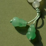 Close-up of a beaded necklace or bracelet with white round beads and two larger green beads on knotted green cords, placed on an olive green surface.