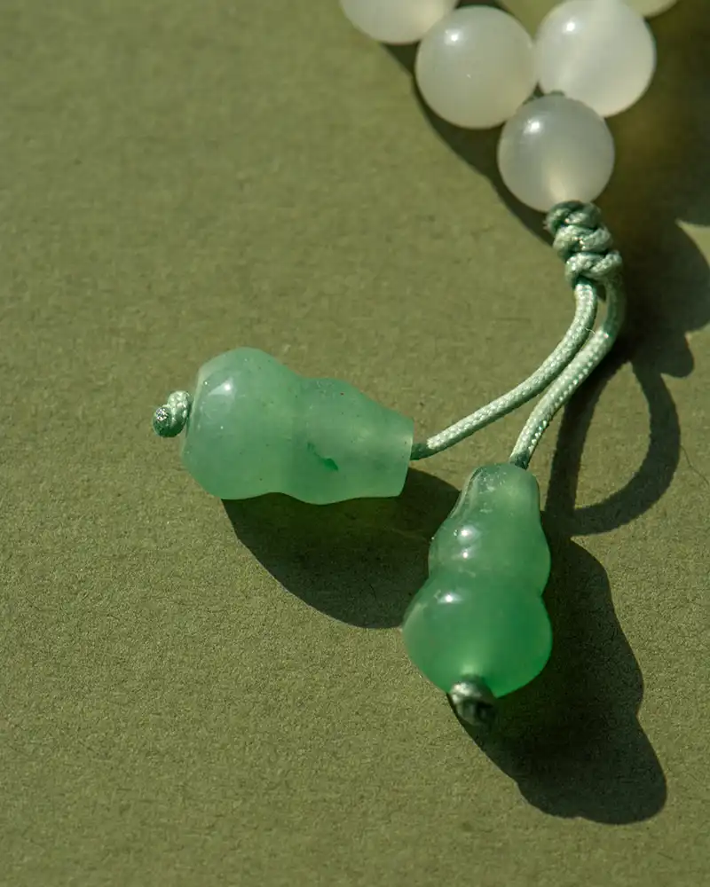 Close-up of a beaded necklace or bracelet with white round beads and two larger green beads on knotted green cords, placed on an olive green surface.