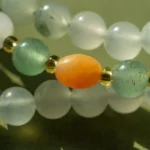 Close-up of a beaded bracelet with frosted white, translucent green, and gold beads, featuring a prominent oval-shaped orange bead in the center, all resting on a green surface.