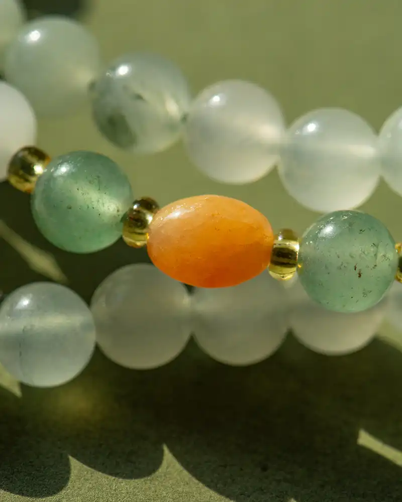 Close-up of a beaded bracelet with frosted white, translucent green, and gold beads, featuring a prominent oval-shaped orange bead in the center, all resting on a green surface.